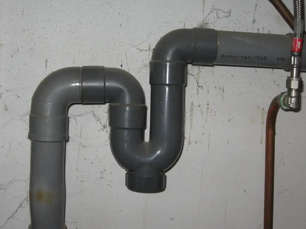 Sifón de PVC gris en forma de curva bajo un fregadero, con el tapón de registro en la parte baja